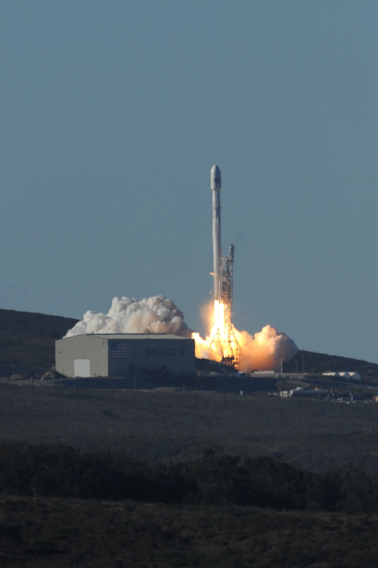 A Falcon 9 rises beside its launch tower in bright flame and white exhaust above the Vandenberg hills while the enclosed CASSIOPE rideshare payloads remain inside the fairing.