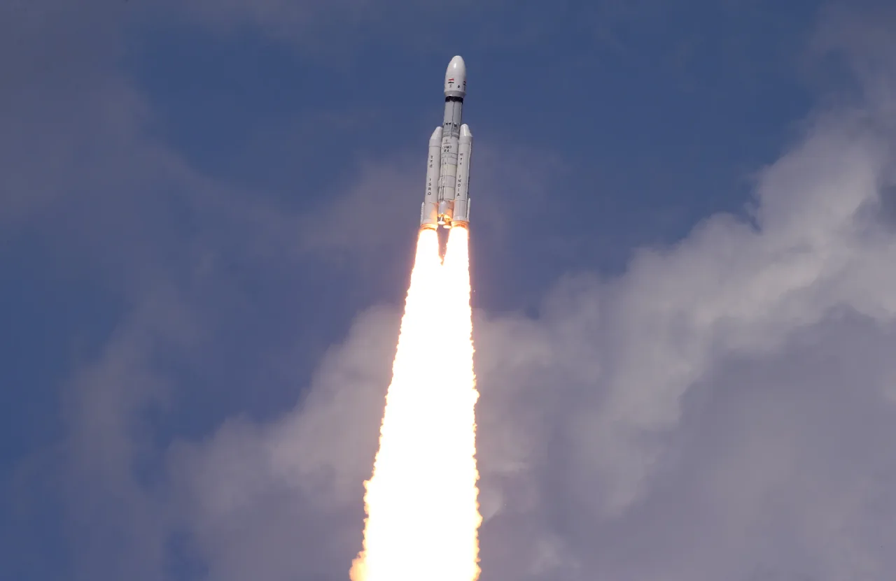 LVM3-M4 climbs against a blue sky with its paired boosters and bright exhaust visible