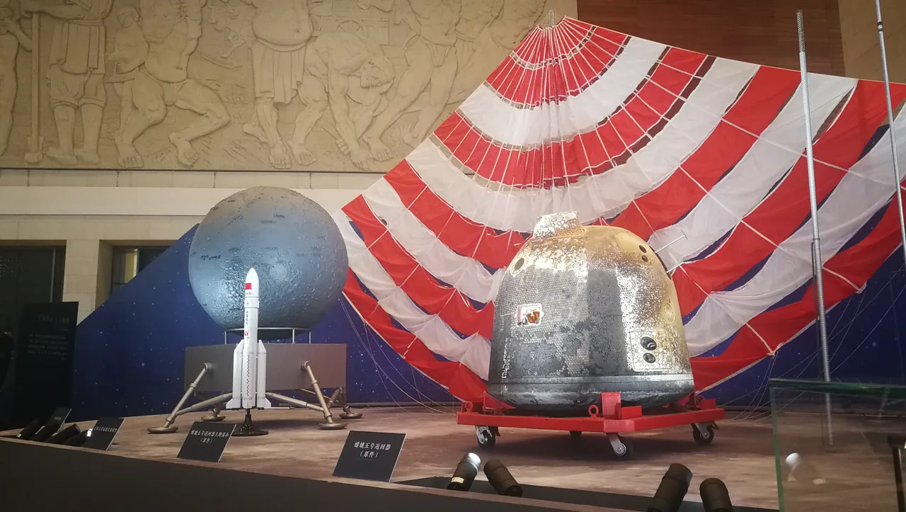 Chang’e-5 return capsule on a red support in a museum, with a red-and-white parachute behind it and smaller display models to the left.