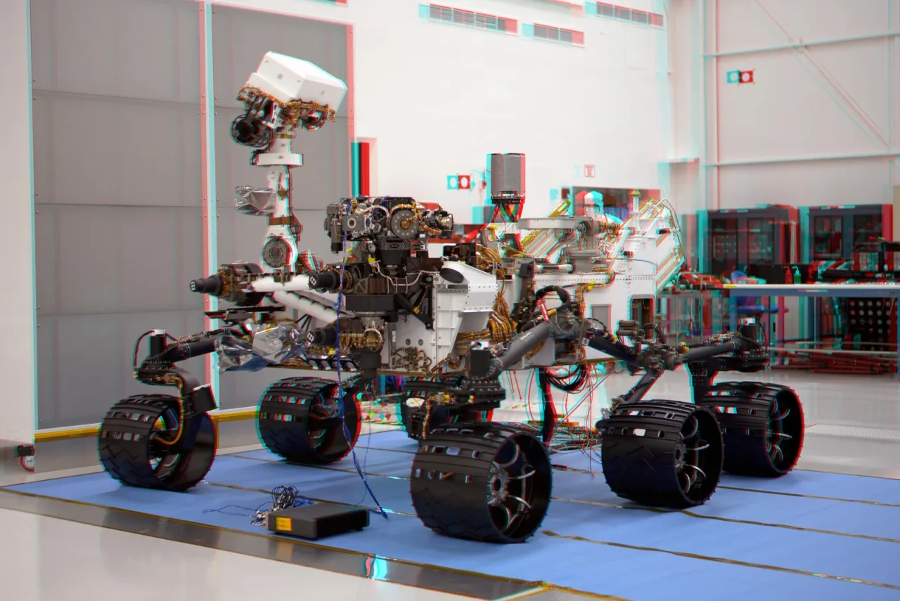 Curiosity stands on six wheels inside an assembly facility, with red and cyan offsets around the mast and equipment