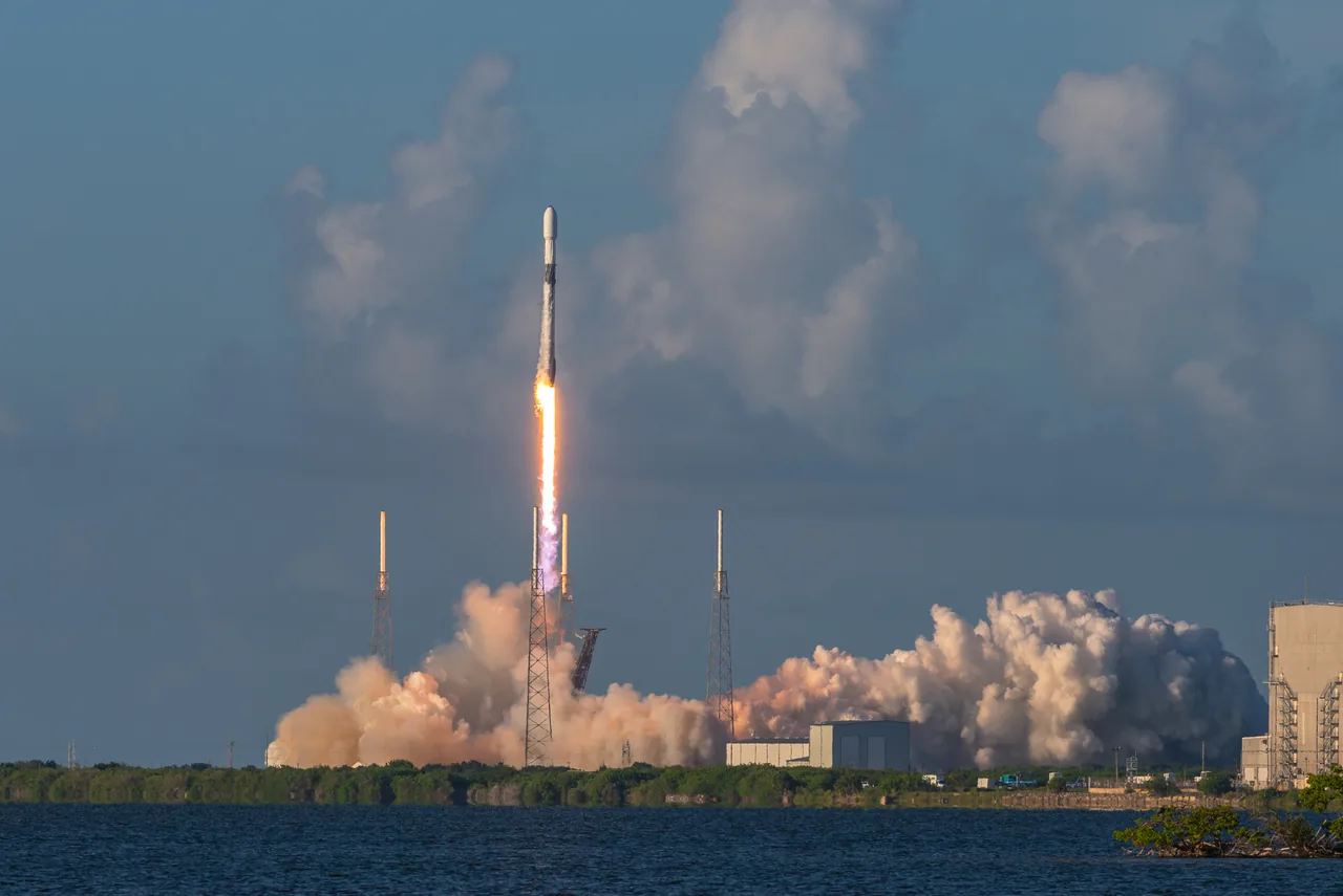 A Falcon 9 climbs above the coast with a bright exhaust plume beneath a blue-gray sky