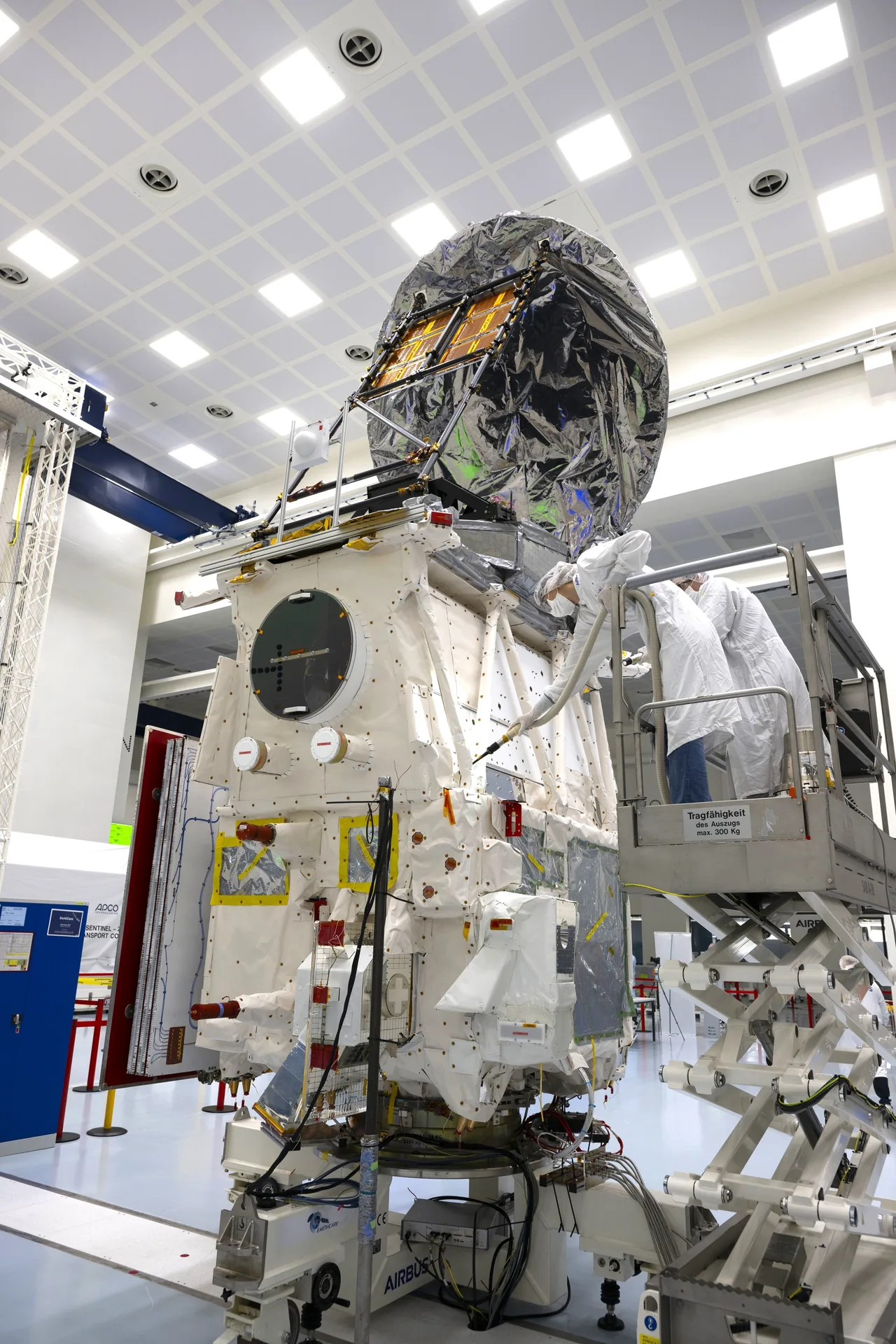 Engineers in cleanroom clothing work beside the tall EarthCARE spacecraft, whose upper structure is covered in reflective silver-colored wrapping above light-colored panels.