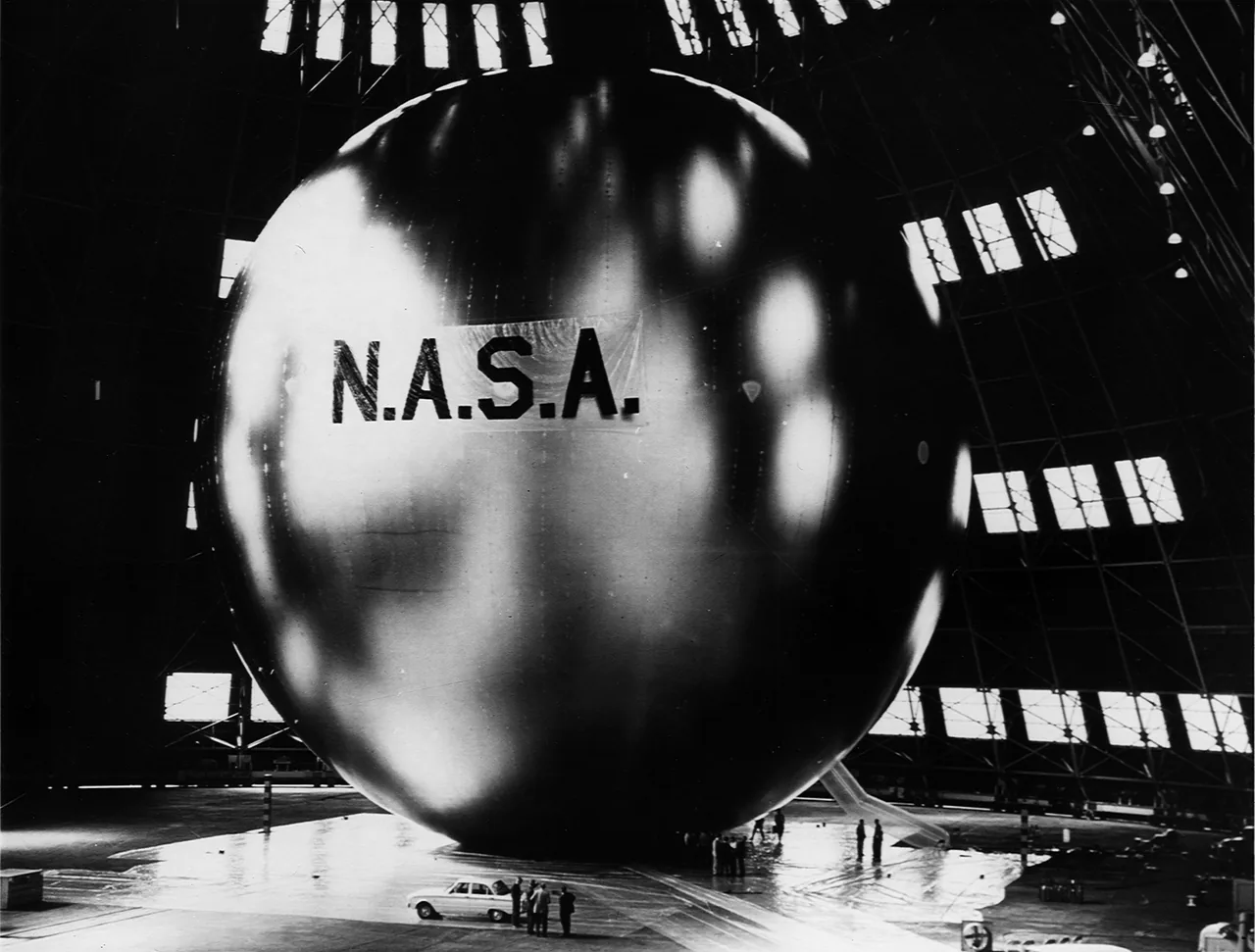 An enormous reflective Echo balloon fills a hangar while people and a car establish its scale