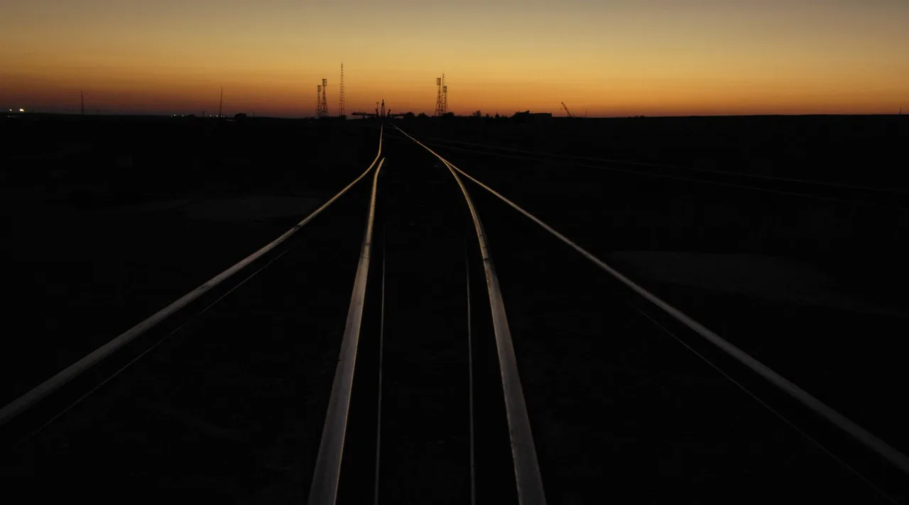Rail tracks converge toward a dark launch complex beneath an orange dawn sky
