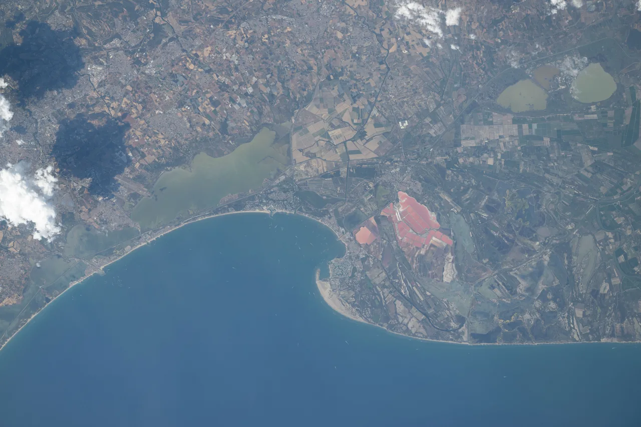 An Italian coastline photographed from orbit, with blue water, fields, settlements, pale lagoons, and scattered clouds