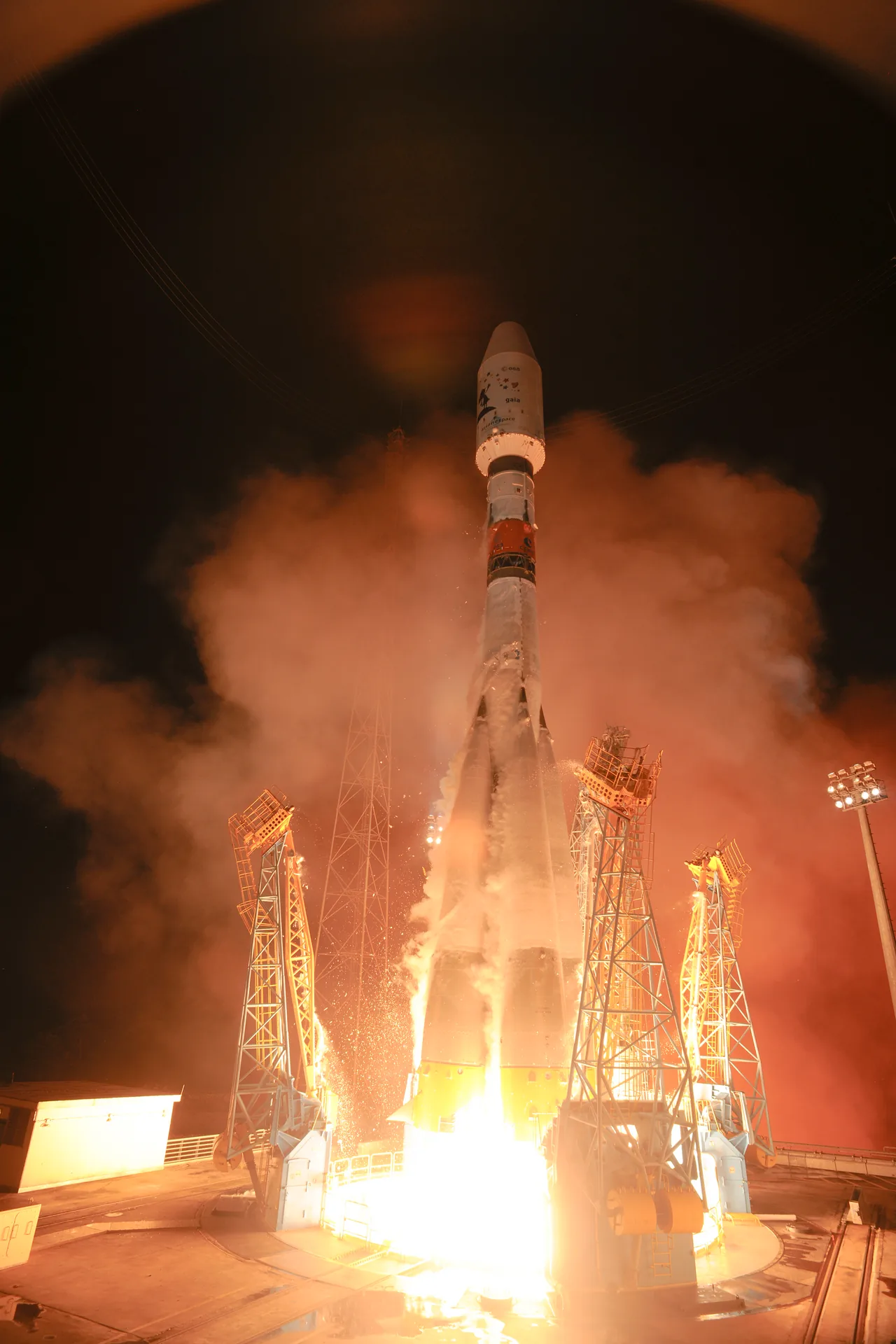 Soyuz lifts off at night carrying Gaia, with a bright plume below a pale rocket marked with the mission name.