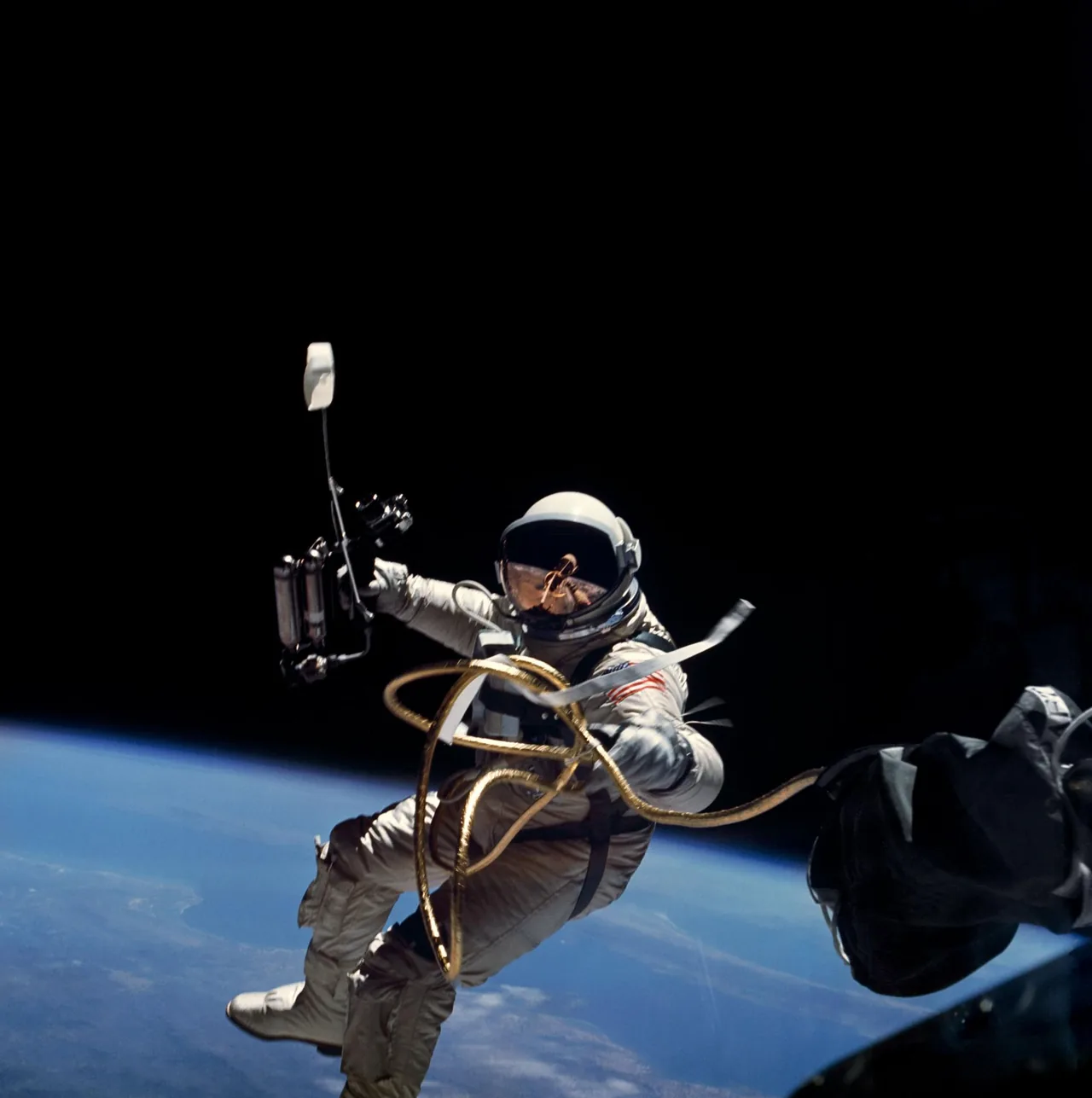 Ed White holds a hand-held maneuvering unit outside Gemini 4, with gold-wrapped connecting lines and Earth below.