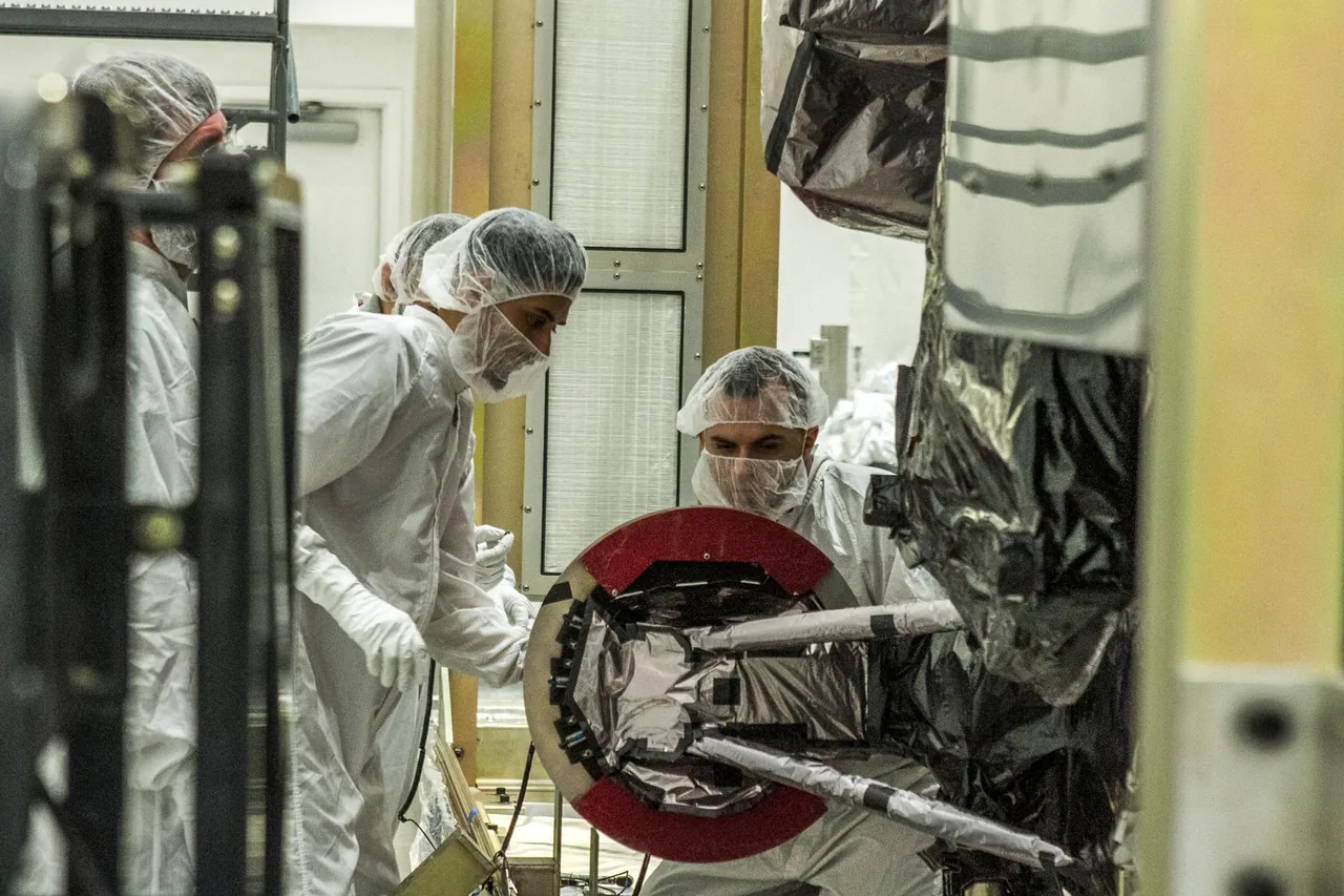 Engineers wearing white cleanroom clothing inspect silver-covered spacecraft hardware beside an open container