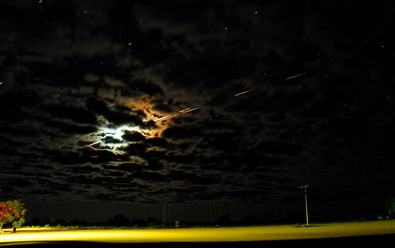 Long bright trails cross a cloudy night sky above a lit field and utility poles during Hayabusa’s artificial reentry.