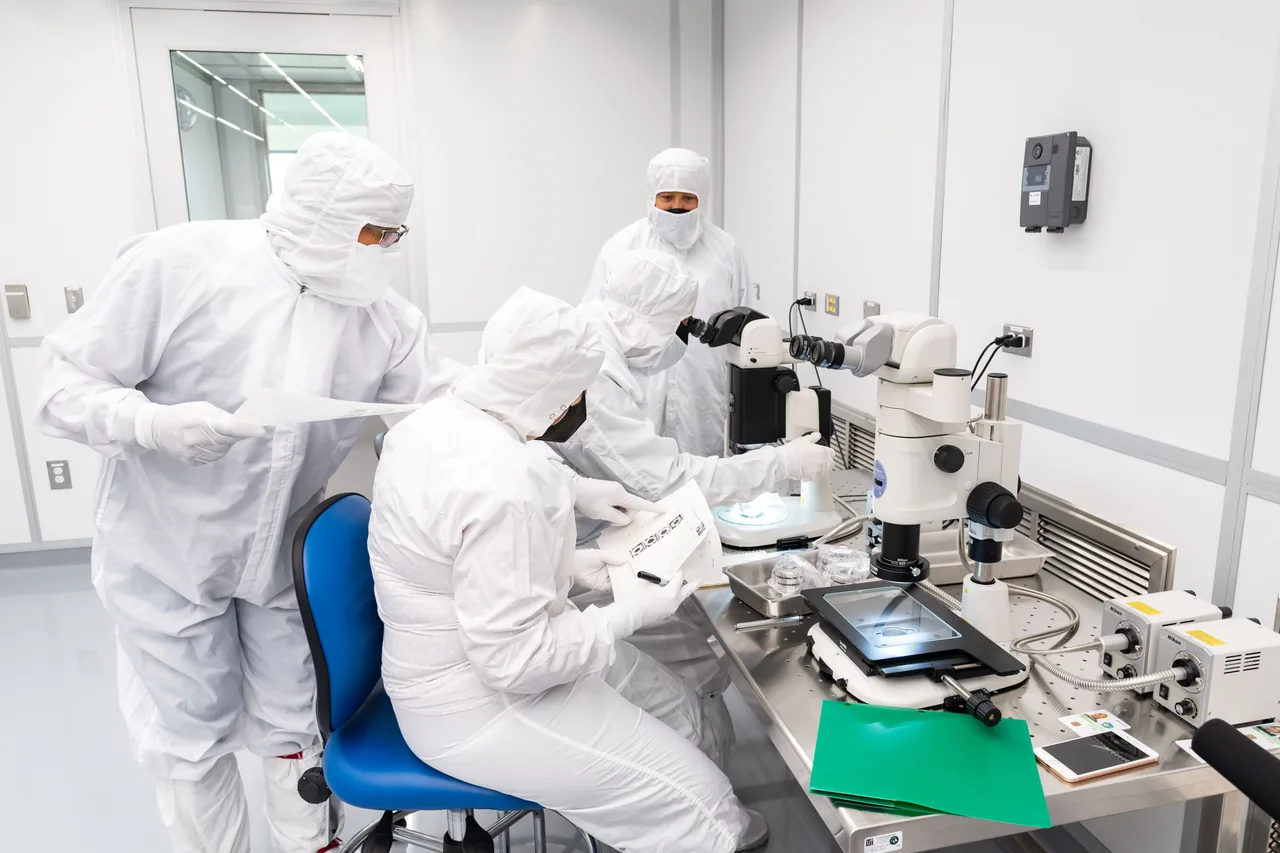 Three people wearing white cleanroom clothing work beside microscopes and sample-handling equipment in a bright laboratory.