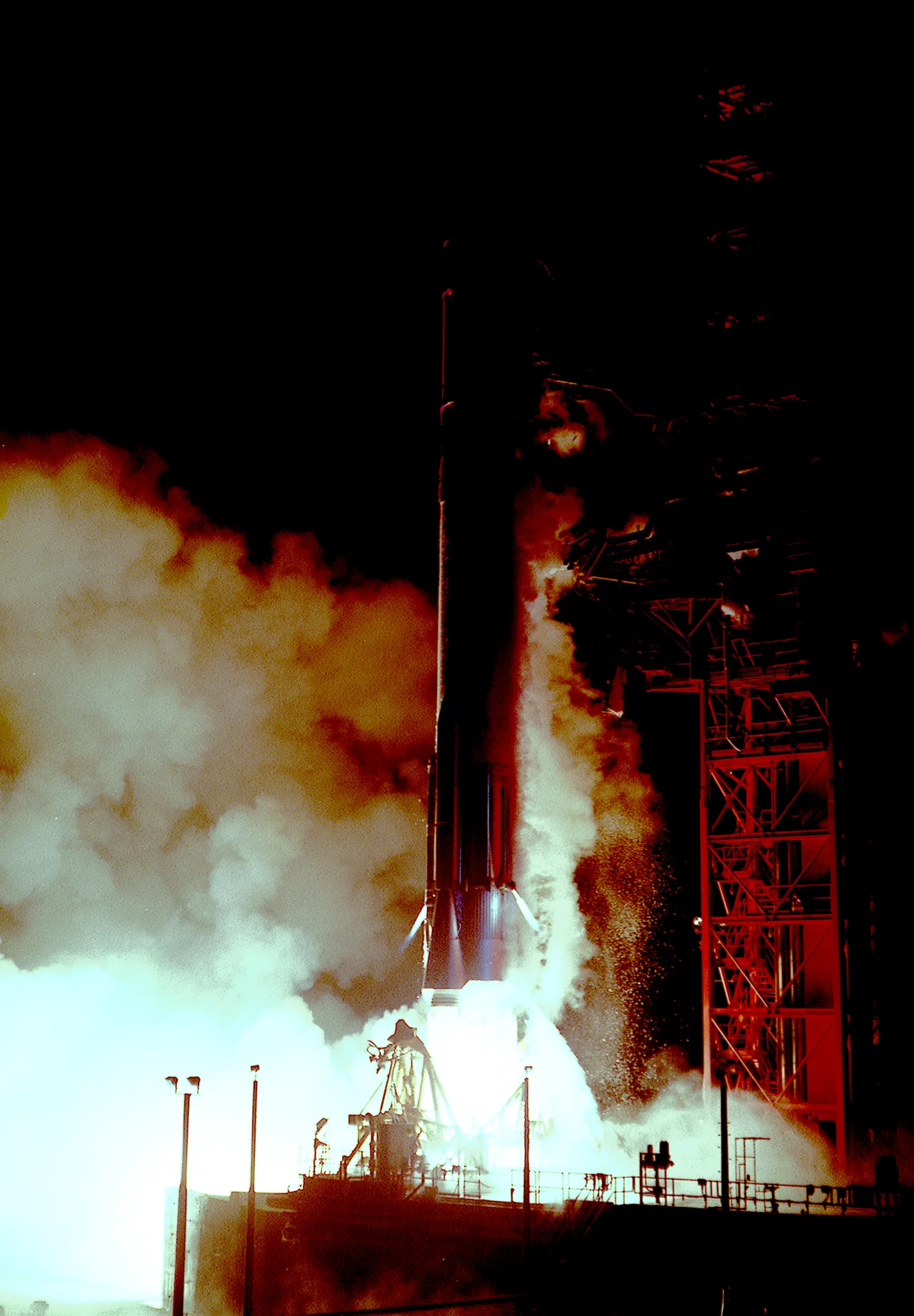 A rocket rises beside a launch tower at night amid bright exhaust and billowing smoke
