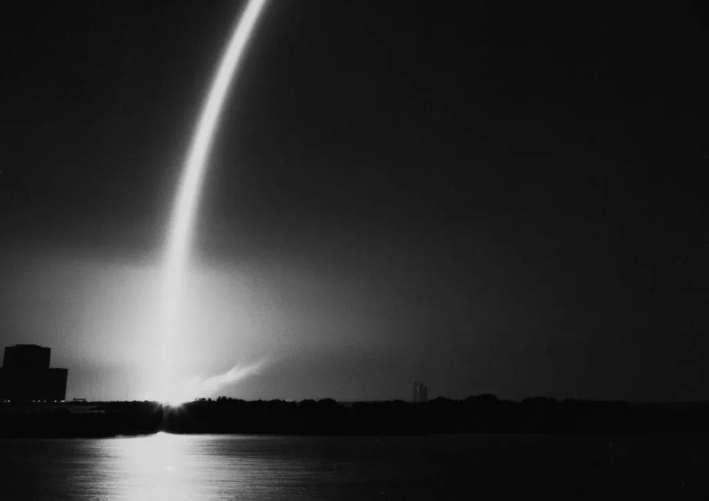 A bright launch trail curves into a dark sky above a low horizon and reflective water