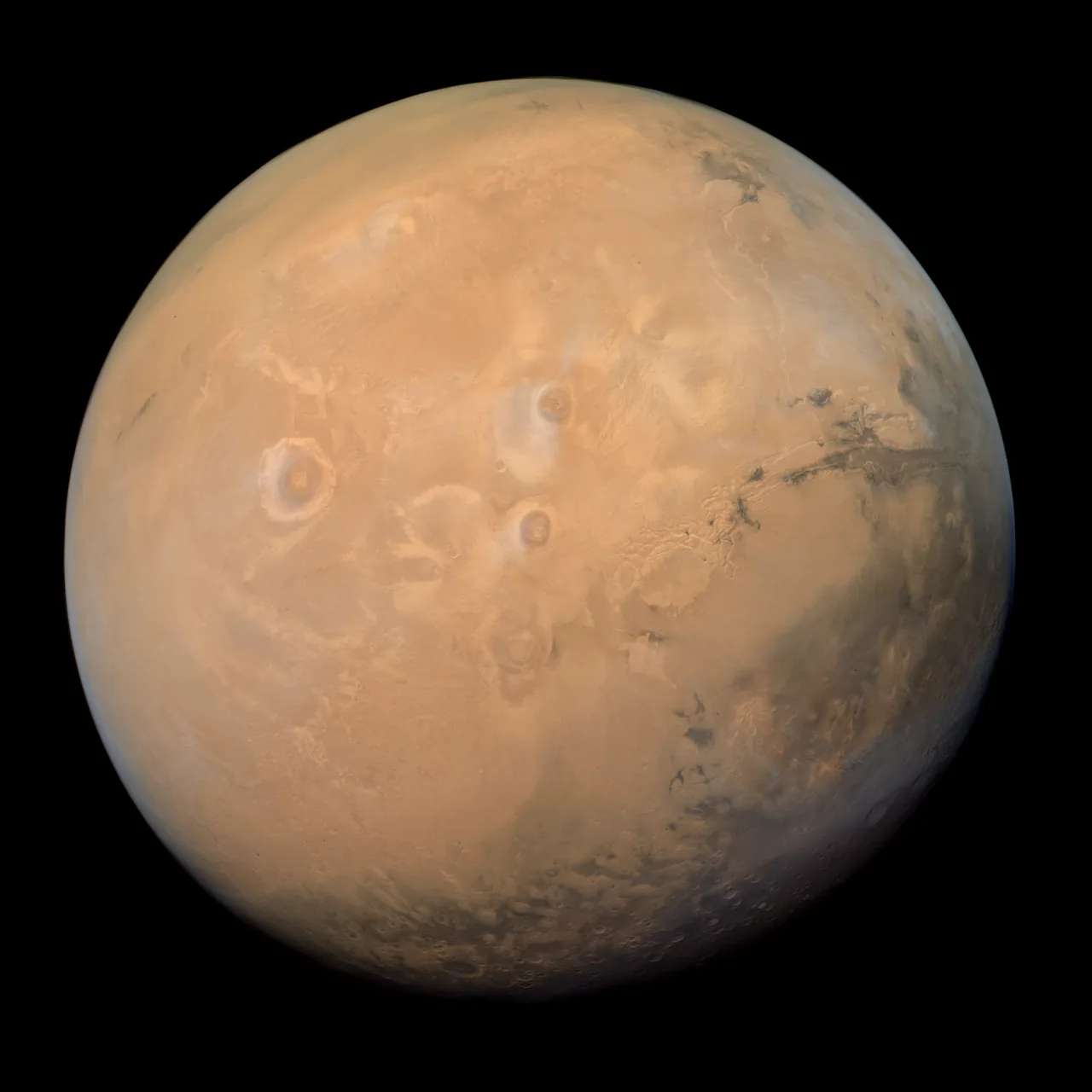 A tan Martian disk shows dark markings, large circular volcanic forms, a long canyon region, and a hazy curved limb.