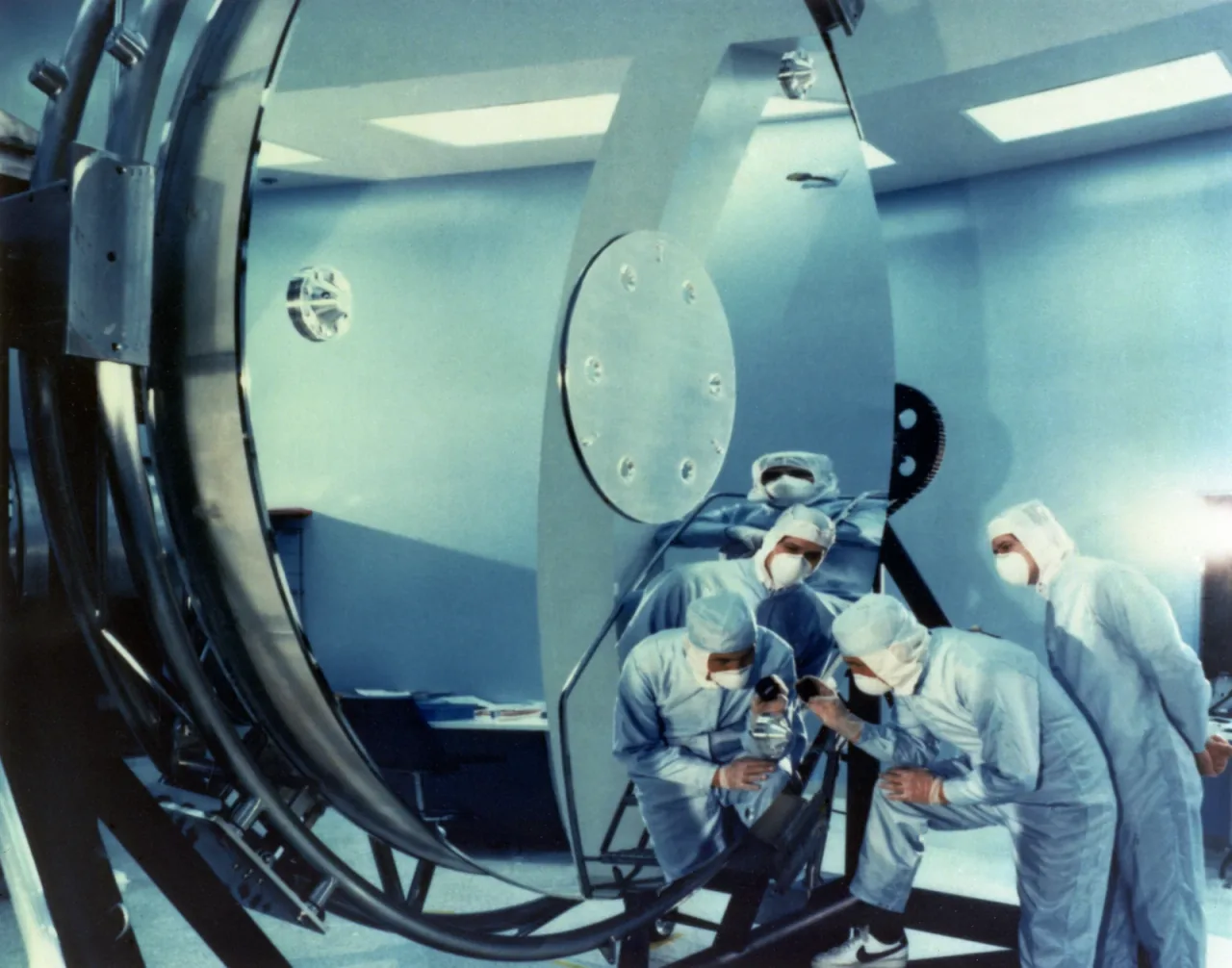 Technicians in pale cleanroom clothing examine a large circular telescope mirror and its reflected surroundings during ground preparation.