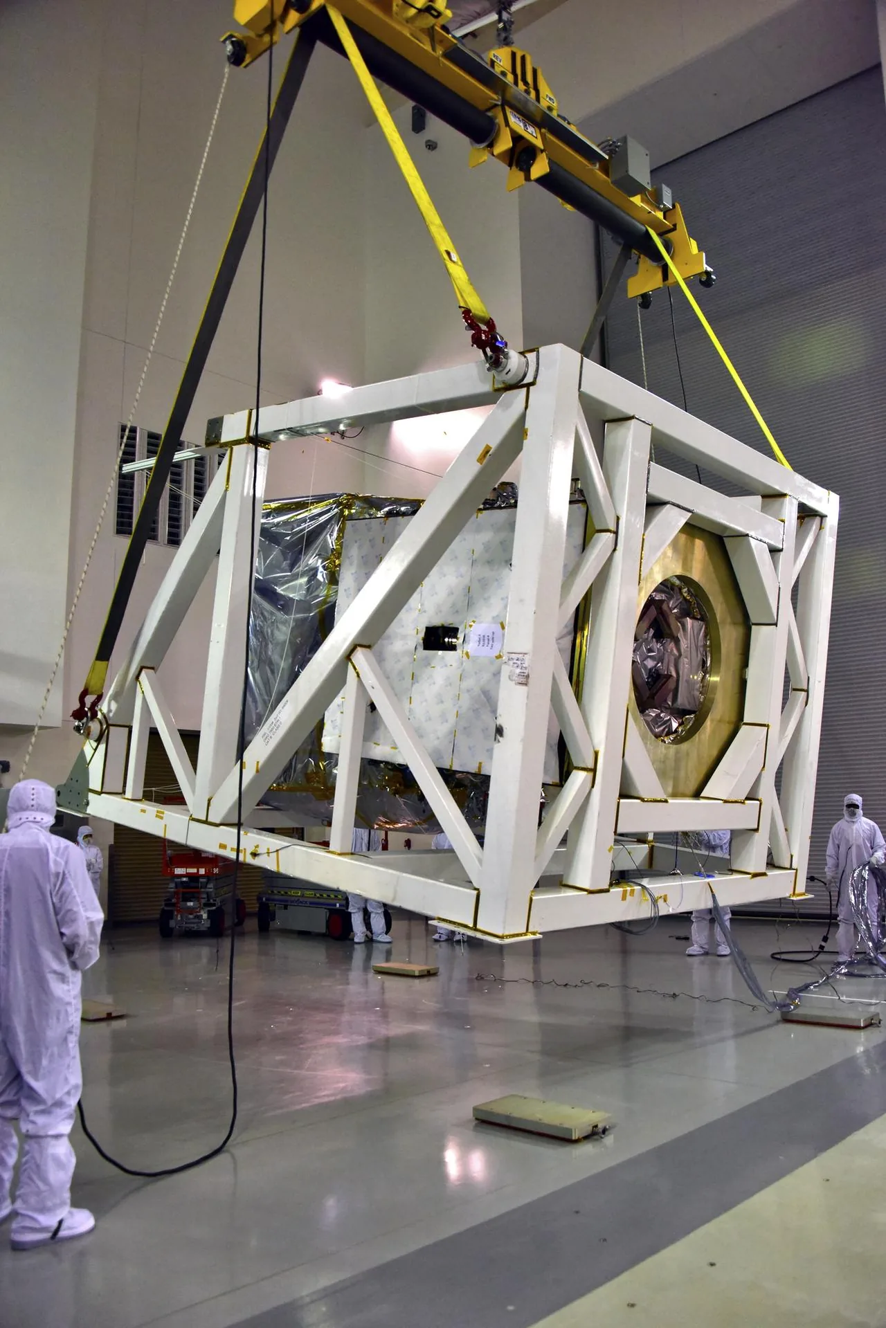 A white support fixture and the foil-covered ICESat-2 spacecraft hang from a yellow hoist while technicians in cleanroom clothing stand nearby.