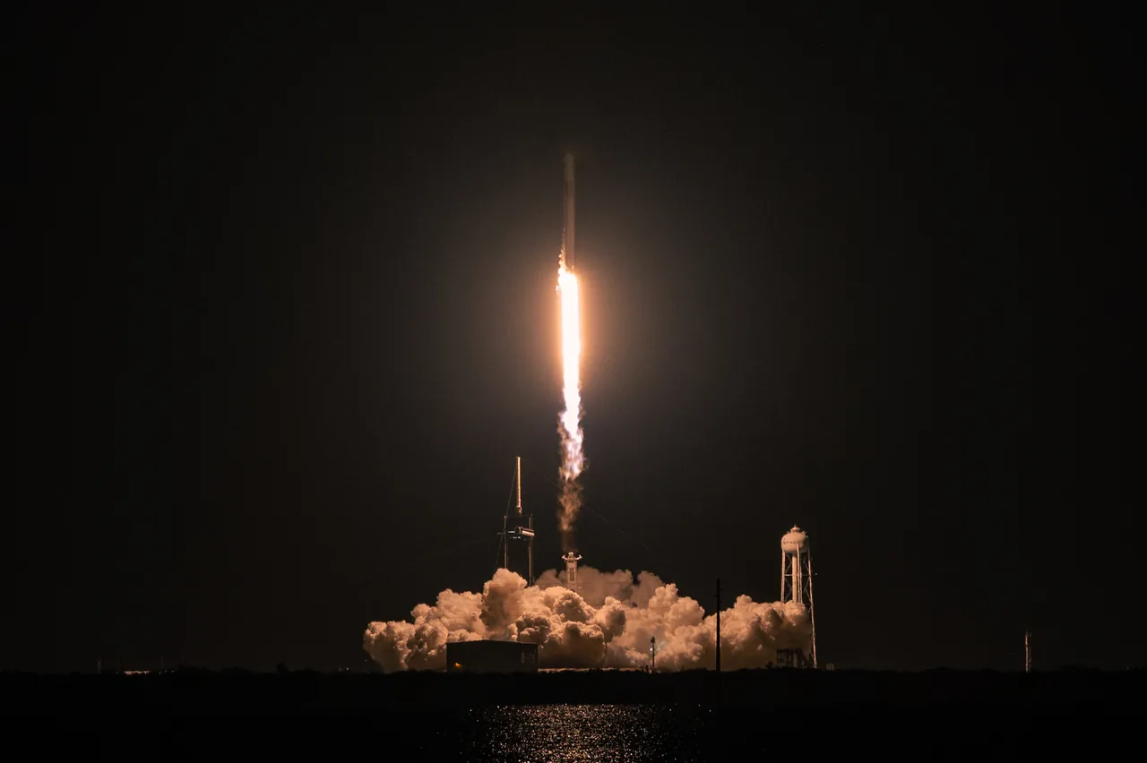 Falcon 9 climbs at night over a launch pad and water reflecting the exhaust