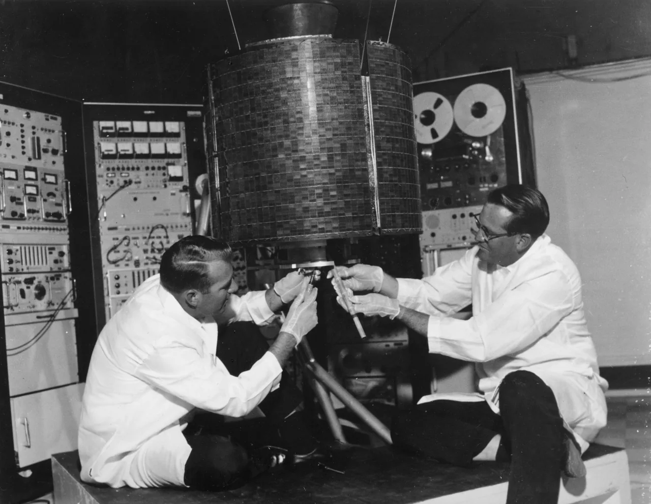Two engineers work beneath the cylindrical Intelsat I spacecraft beside racks of ground equipment