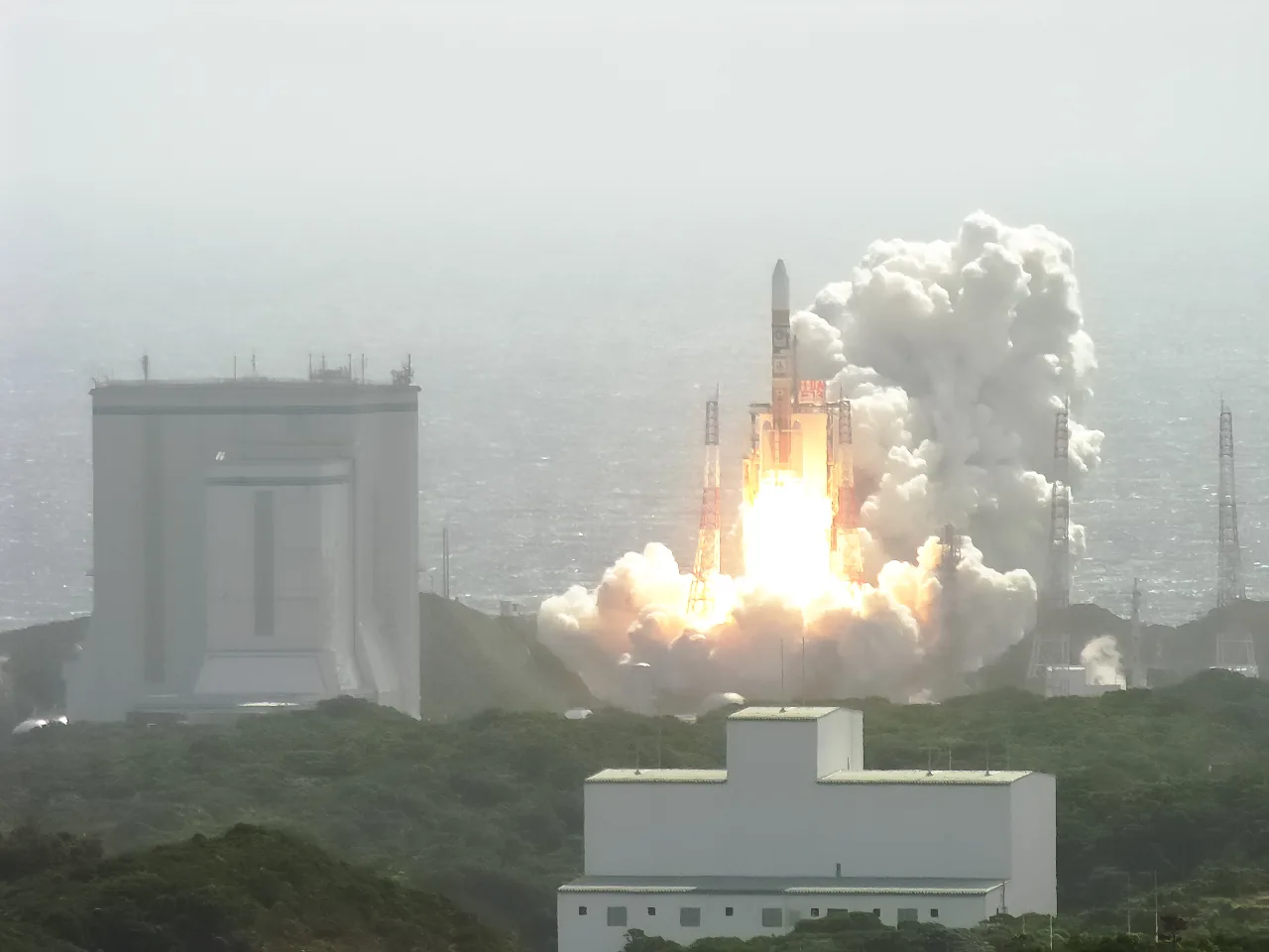 H-IIA Flight 13 rises from its coastal launch pad carrying Kaguya, with a bright exhaust plume, rolling white clouds and launch-site buildings in the foreground.
