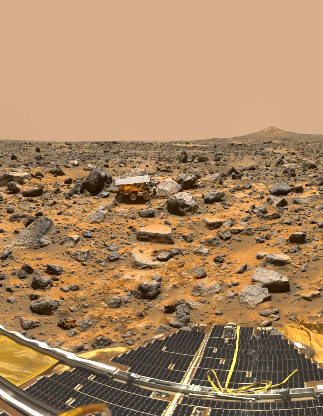 Sojourner stands beside a pale rock on a boulder-strewn plain, with the Pathfinder lander’s solar panels in the foreground.