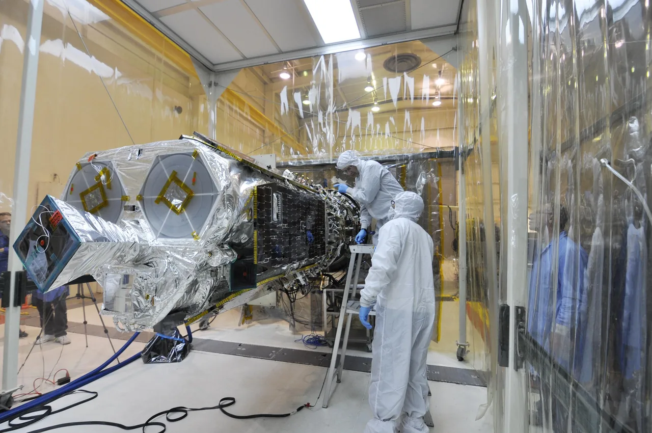 Technicians in cleanroom clothing work beside the silver-covered NuSTAR spacecraft and its rocket connection inside a transparent enclosure.