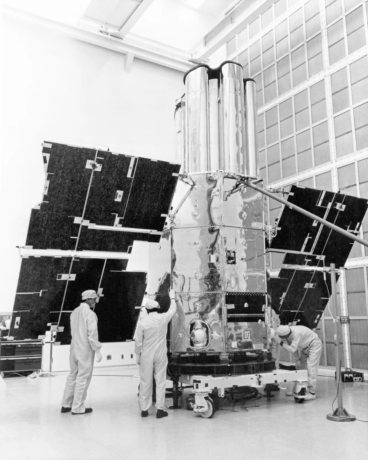 Technicians check a tall cylindrical OAO-3 spacecraft with rectangular solar panels in a ground facility