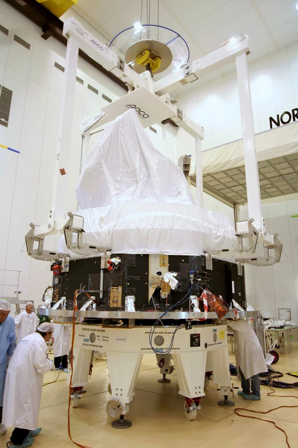 Planck stands on a support platform among cleanroom technicians, with a white protective covering over its upper payload and lifting equipment above.