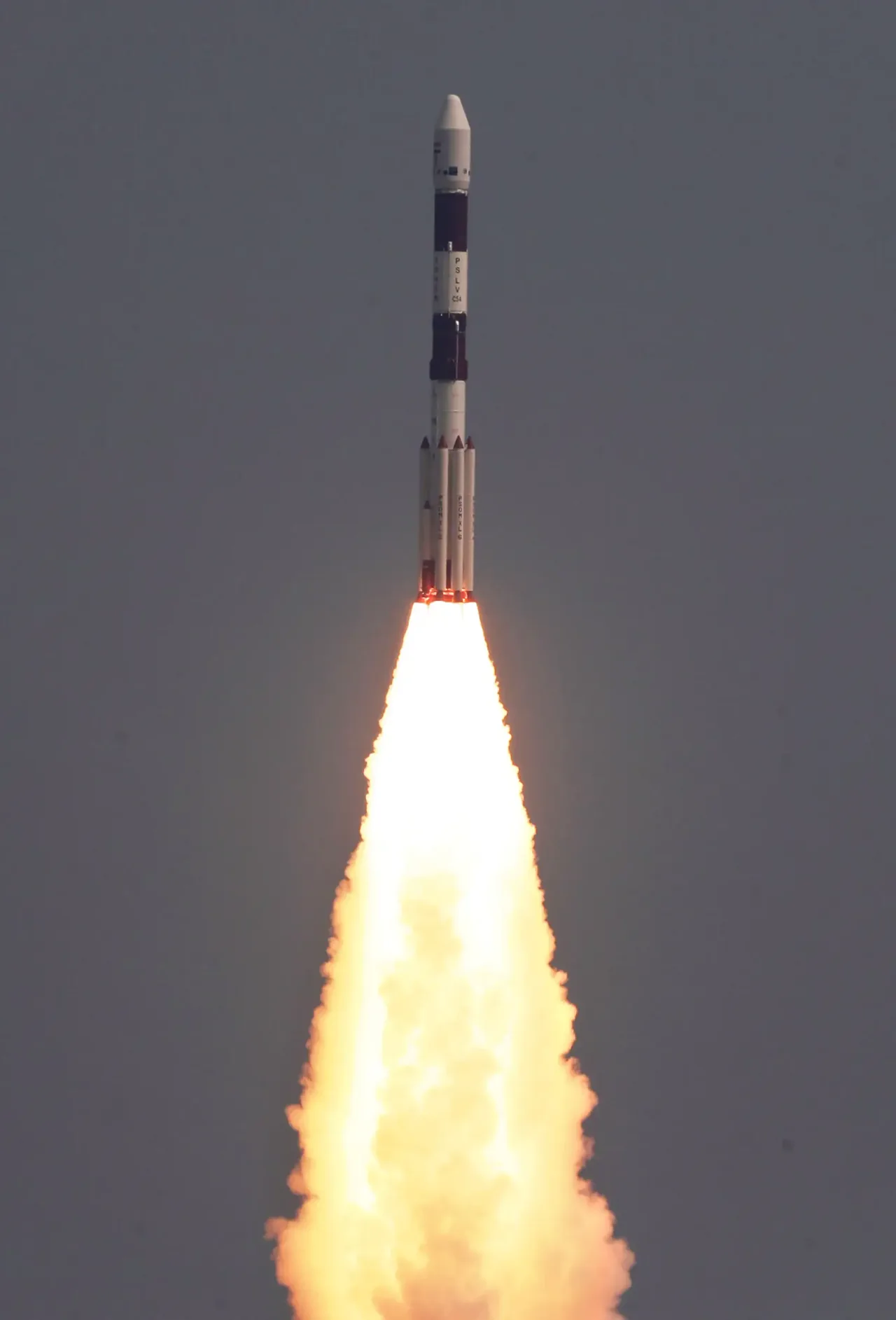 A white PSLV rocket rises through a gray sky with clustered boosters and a bright exhaust plume