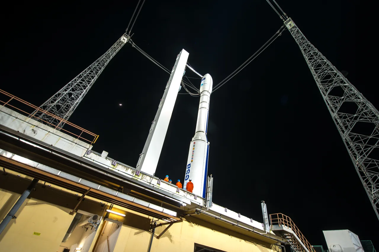 A white Vega rocket stands between lattice towers at night, with workers near its base