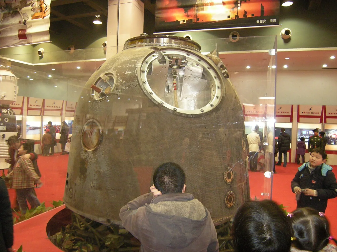 Visitors stand around Shenzhou 7’s dark reentry capsule in a brightly lit exhibition hall