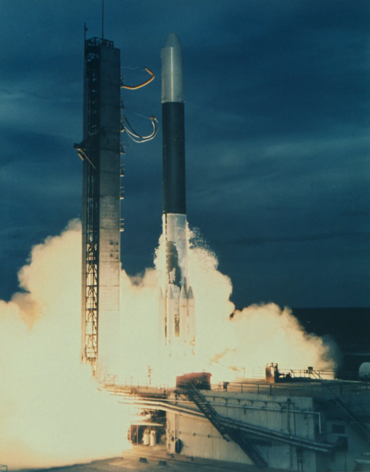 A rocket lifts from a launch pad beside a service tower amid a bright exhaust plume