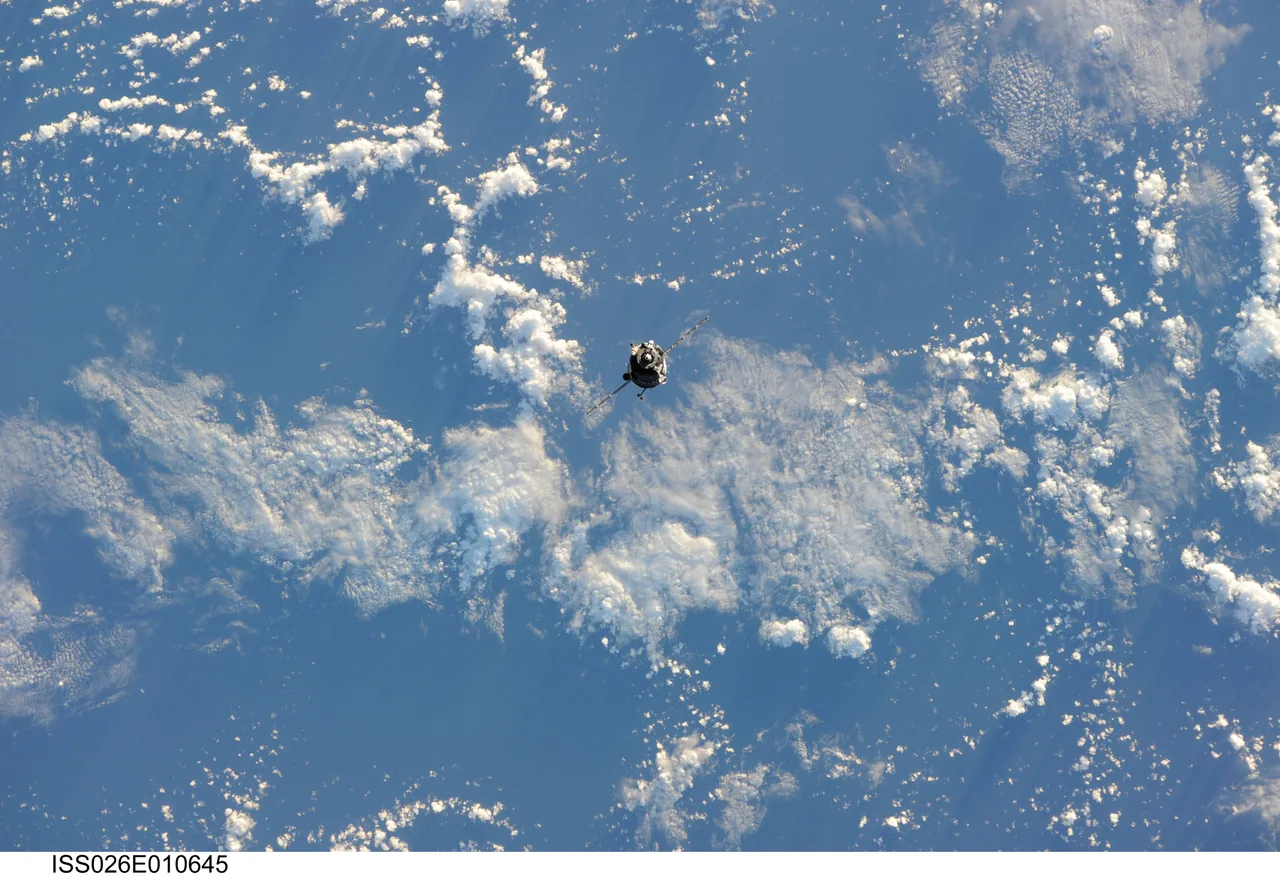 A small Soyuz spacecraft with extended solar panels appears against Earth’s blue ocean and scattered white clouds.
