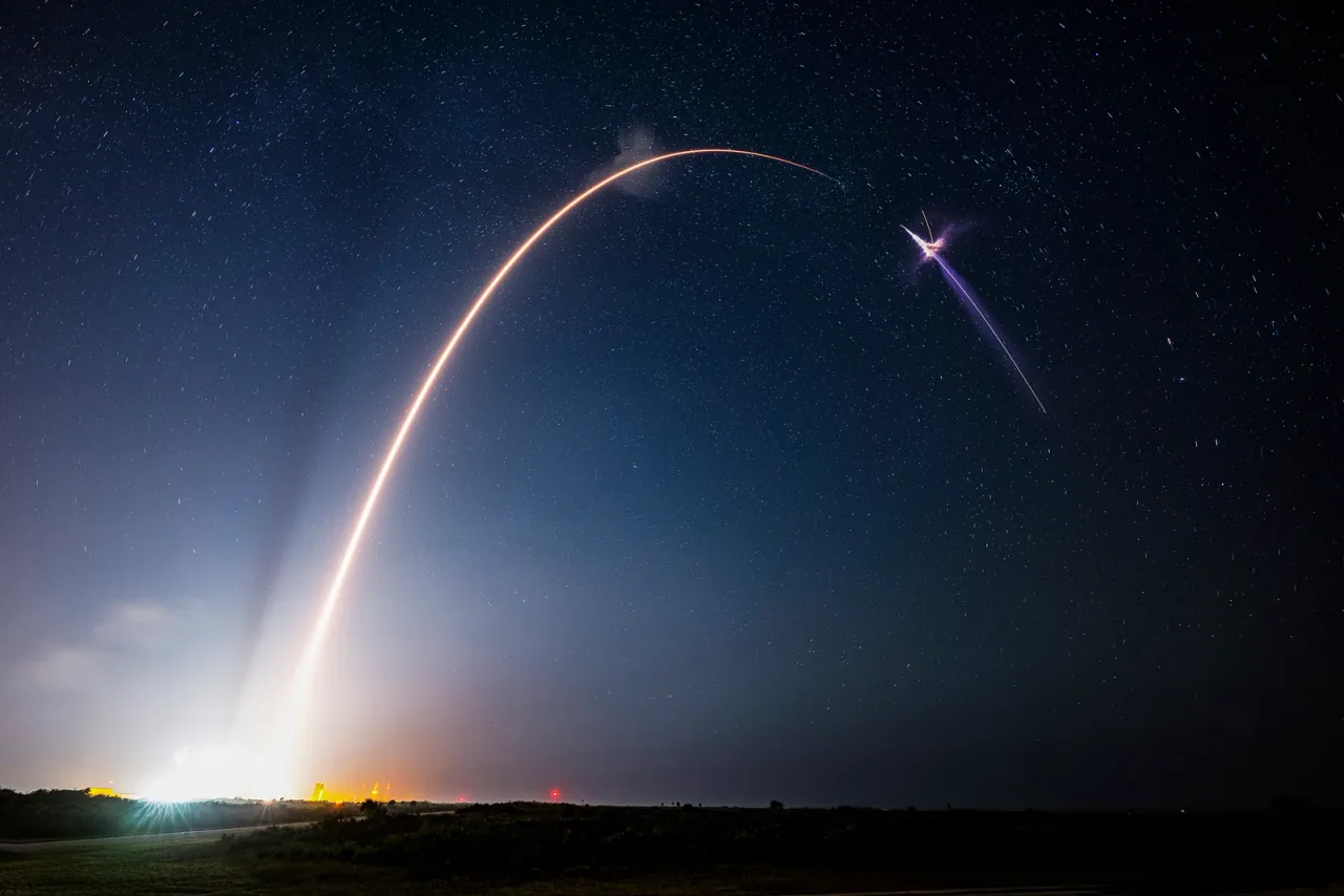 A bright curved launch trail crosses a starry night sky above a low dark horizon