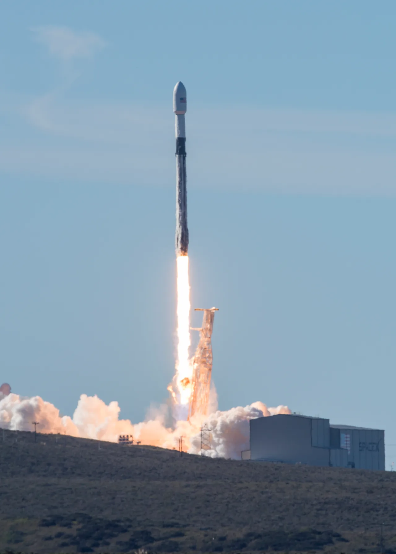 A Falcon 9 carrying SSO-A rises above its launch tower and hillside under a clear blue sky with bright exhaust and smoke below.