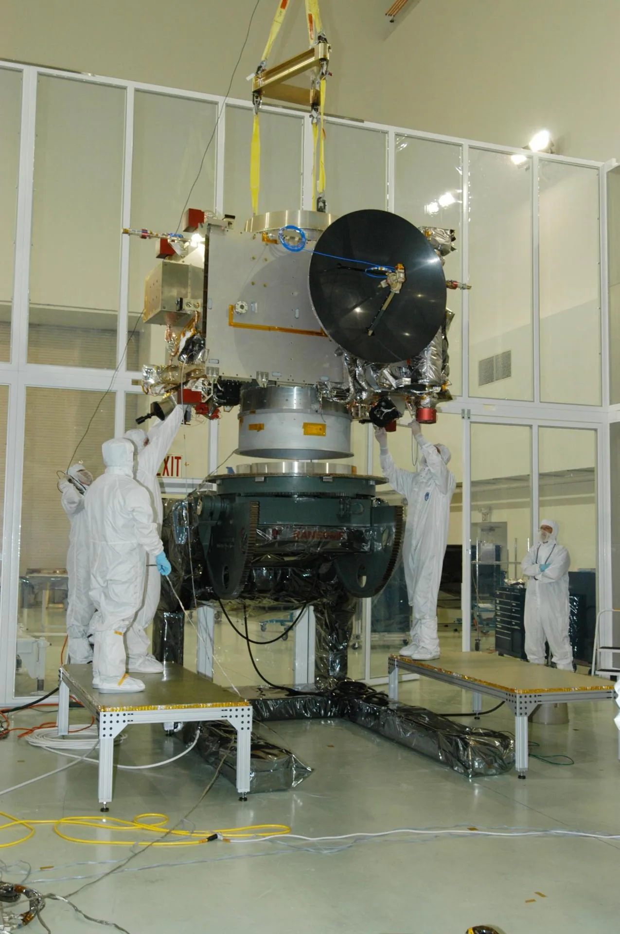 Technicians guide a spacecraft with a large dark dish while lifting equipment supports it