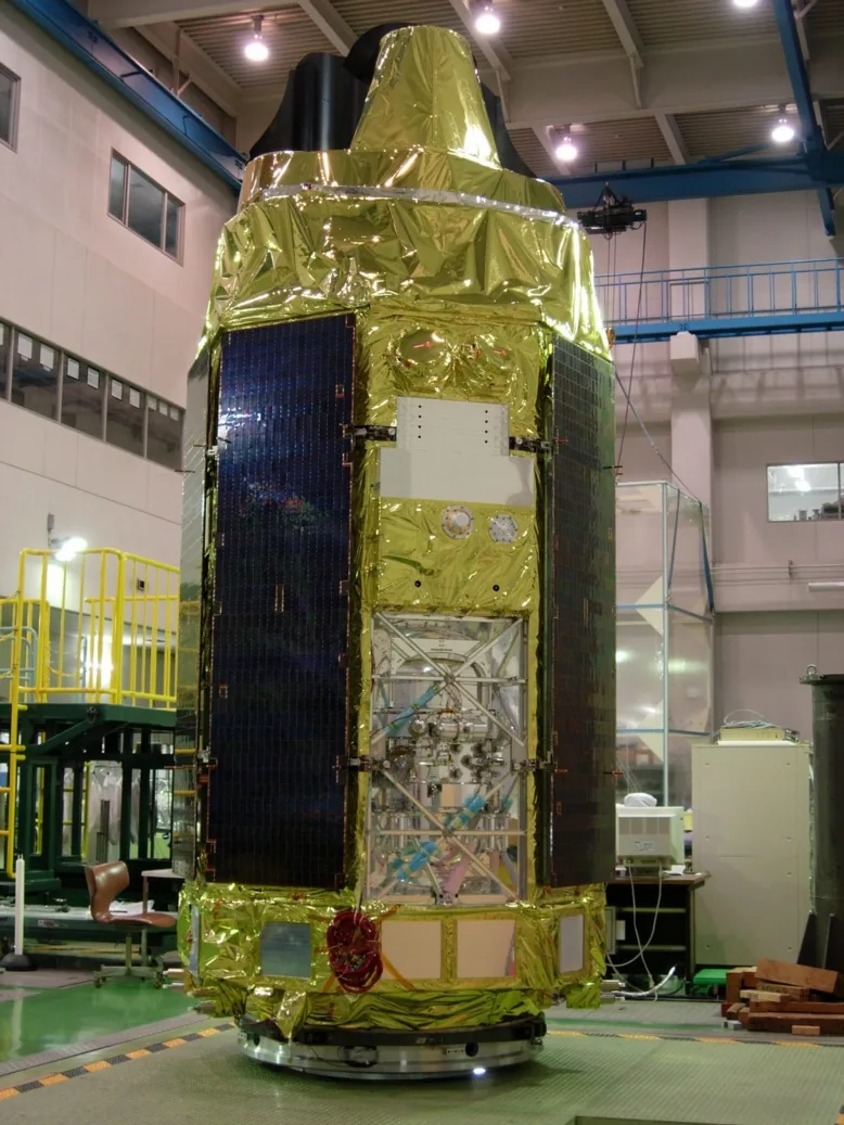 The tall Suzaku spacecraft stands in a ground facility with gold thermal covering and dark solar panels