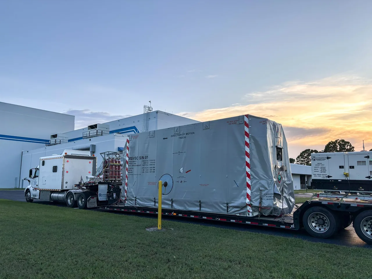 A covered spacecraft shipping container sits on a truck outside a processing facility near sunset