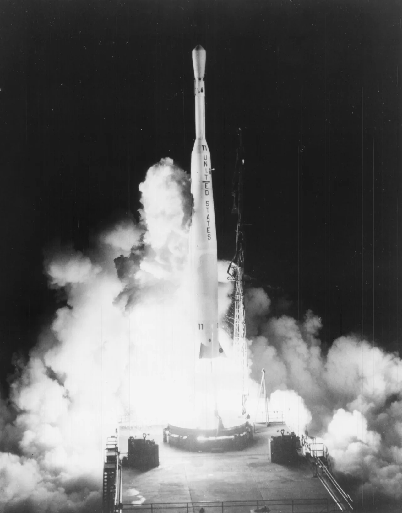Black-and-white photograph of a Thor-Delta rocket amid bright exhaust and billowing smoke at its launch pad