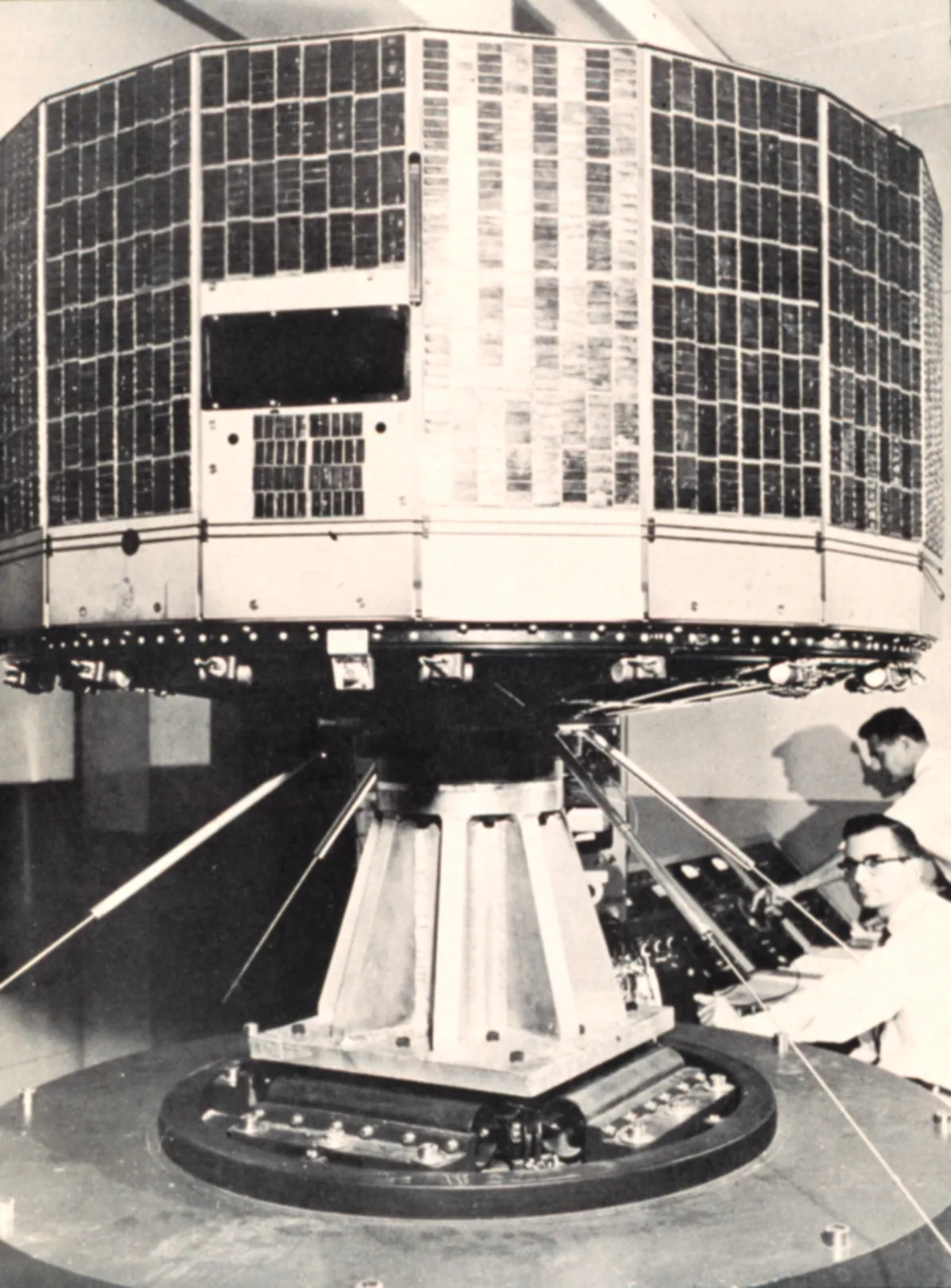 A drum-shaped TIROS-1 satellite covered in solar cells stands on test equipment beside technicians
