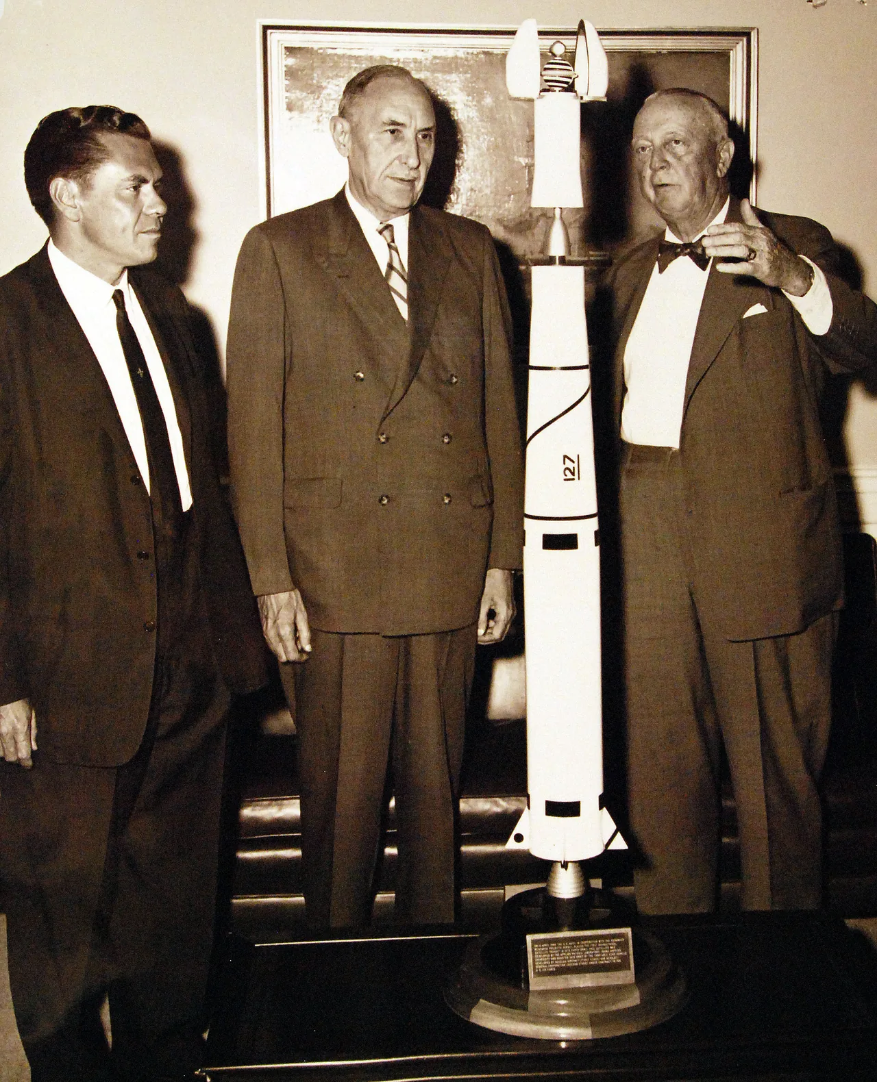 Three officials stand beside a scale model of a launch vehicle and its Transit satellite payload