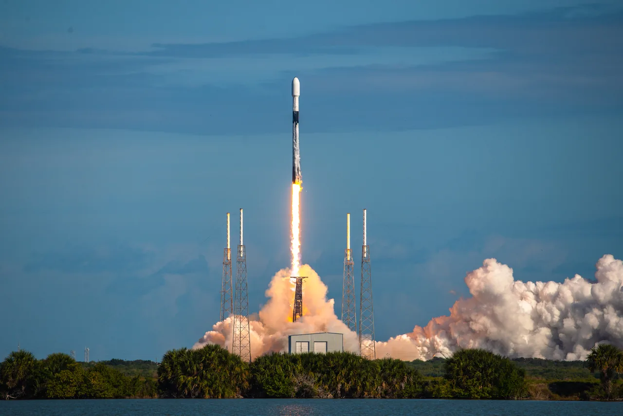 A Falcon 9 rises above Cape Canaveral on a bright exhaust column, with smoke spreading across the launch site beneath a blue sky.