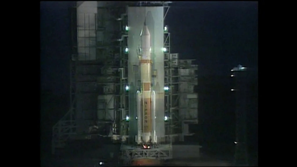 H-II stands beside its launch tower at night, with illumination near the base and black borders around the source frame.