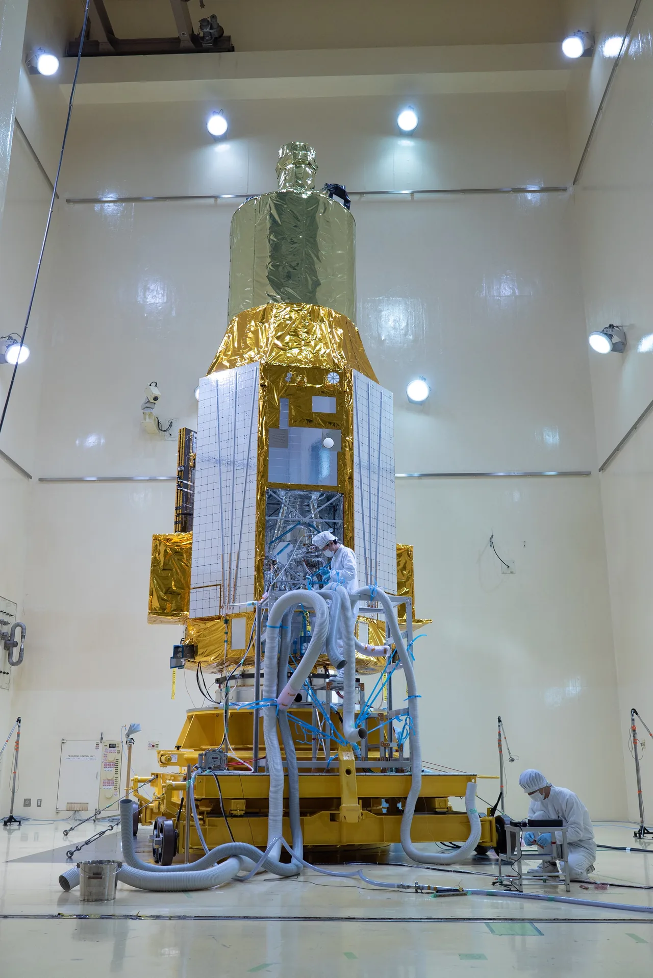 Gold- and silver-covered XRISM spacecraft stands upright in a test room with technicians and service hoses.