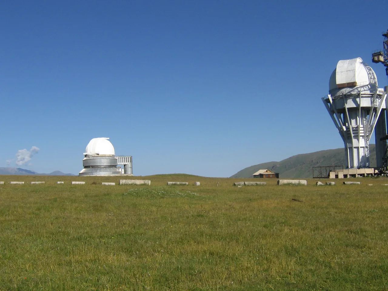 Two white telescope domes at Assy-Turgen Observatory on a grassy plateau beneath a blue sky