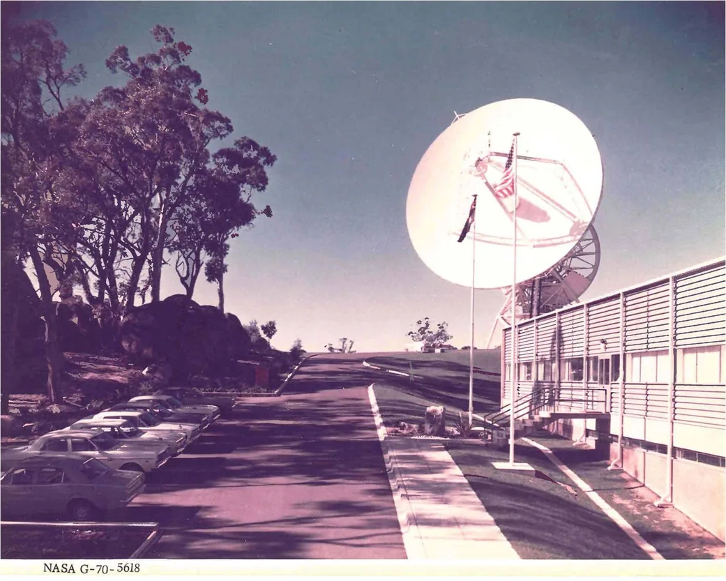 Honeysuckle Creek’s white tracking dish beside an operations building, access road, parked cars and trees