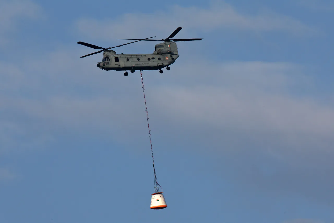 An Indian Air Force Chinook carries a capsule-shaped test article on a long sling against the sky.