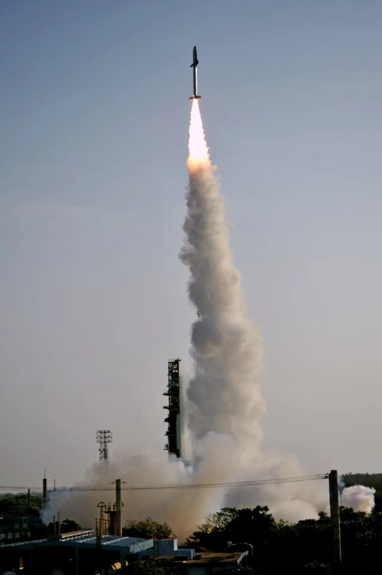 A winged test vehicle sits atop a slender ascending booster with a bright exhaust plume above the launch site.