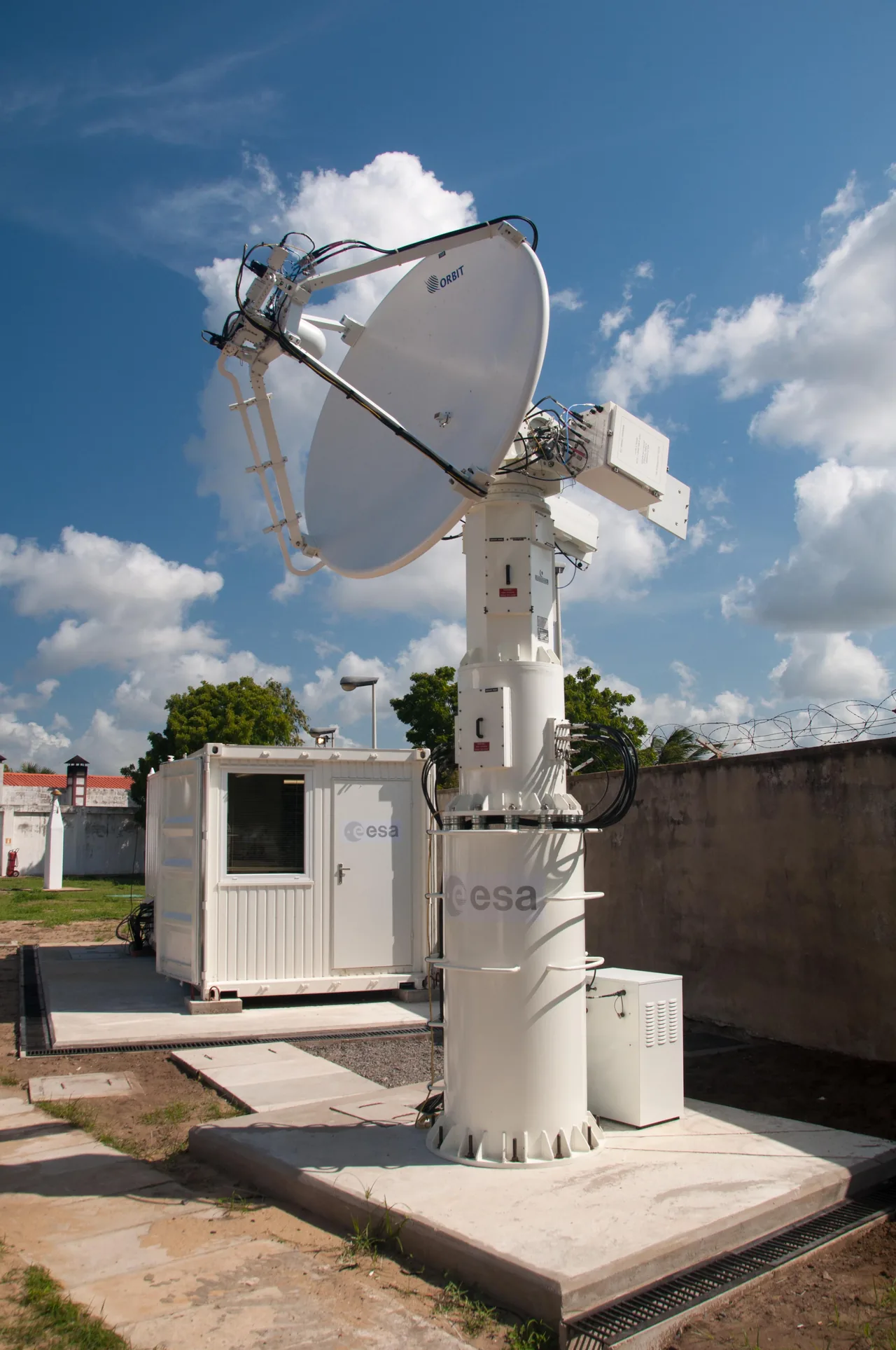 White tracking dish, its support mast and attached cables beside a small equipment cabin under a blue sky