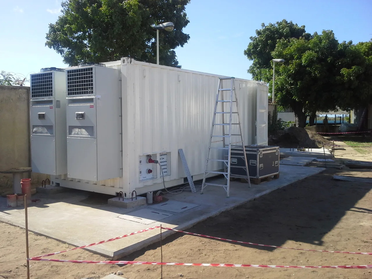 White equipment container with large external cooling units, a ladder and nearby ground works