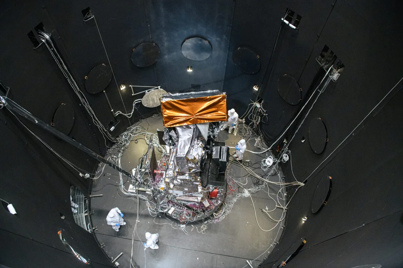 Overhead view of the foil-covered PACE observatory, cables and cleanroom-clad technicians inside a large dark-walled test chamber