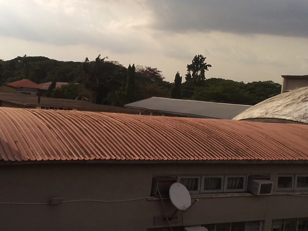 A pale dish antenna stands below a red corrugated roof, with trees and cloudy sky behind the buildings
